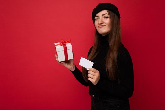 Attractive positive young brunette woman wearing black sweater and hat isolated on red background holding credit card and white gift box with red ribbon looking at camera.