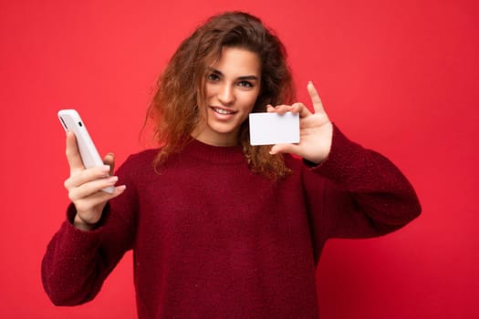 Young smiling beautiful curly brunette woman with sincere emotions poising isolated over background wall with empty space wearing casual dark red sweater holding credit card and using mobile phone . Internet banking concept.