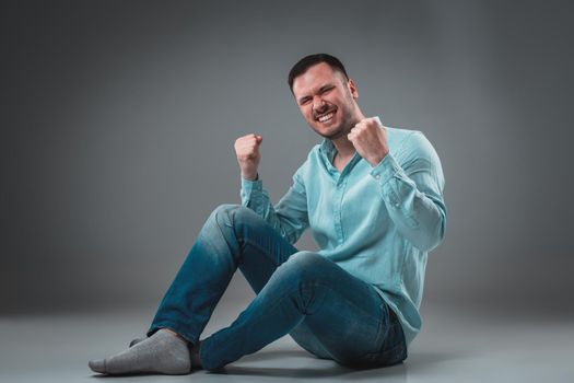 Handsome young man sitting on a floor with raised hands gesturing happiness on gray background. A man in jeans and a blue shirt