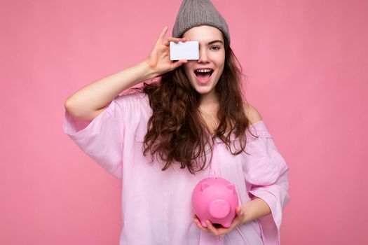 Charming happy joyful young brunette woman wearing shirt isolated on pink background with free space and holding pink pig money box and credit card for mockup.