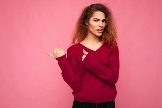 Photo of young shocked beautiful brunette wavy woman with sincere emotions wearing casual pink jersey isolated over pink background with copy space and pointing at free space for advertising.