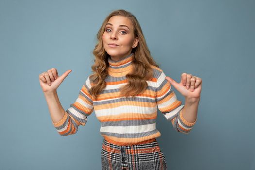 Portrait photo of young pretty beautiful attractive blonde woman with wavy-hair wearing striped sweater isolated over blue background with empty space for text and showing fingers to herself. Me concept.