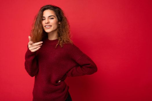 Young positive happy beautiful brunette curly woman with sincere emotions wearing casual red pullover isolated on red background with copy space.