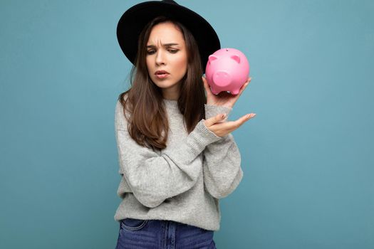 Portrait of thinking angry young beautiful pretty brunette woman wearing gray pullover and black hat isolated over blue background with free space and holding pink piggy box. Money box.