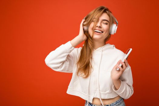 Photo of beautiful happy smiling young blonde woman wearing white hoodie isolated over colourful background wall with copy space for text wearing white wireless bluetooth earphones listening to cool music and holding mobile phone.