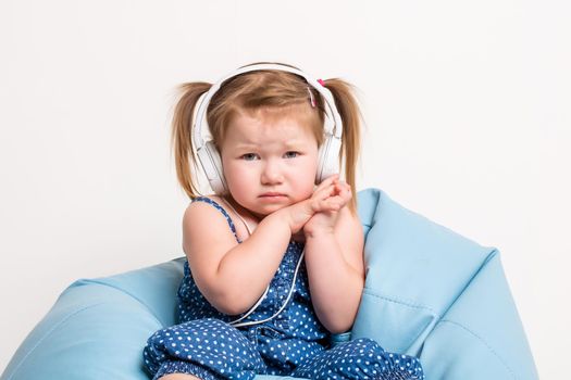 Cute little girl in headphones listening to music using a tablet while sitting on blue big bag. On white background. A child looks at the camera