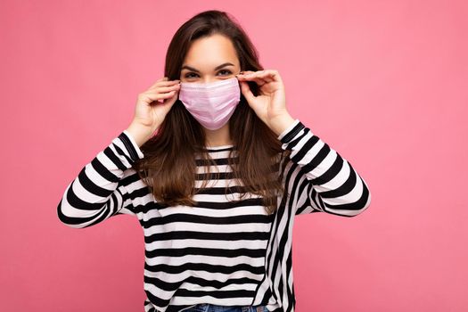 Photo shot of young beautiful brunette woman in reusable virus protective mask on face against coronavirus isolated on the pink background wall with free space for text.