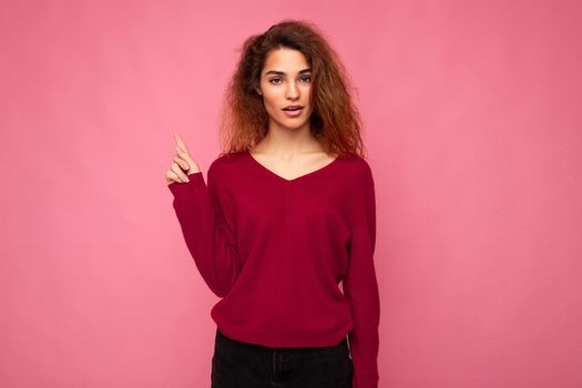Photo of young shocked beautiful brunette wavy woman with sincere emotions wearing casual pink sweater isolated over pink background with copy space and pointing at free space for mockup.