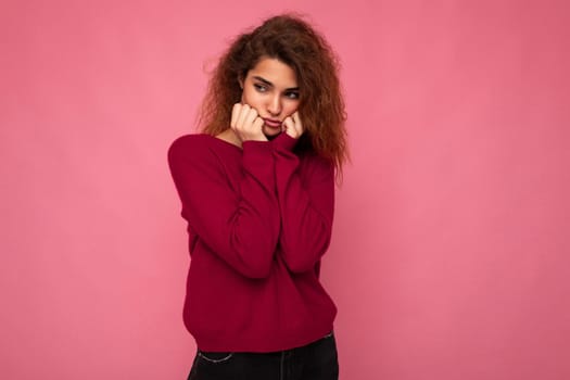 Young emotional offended touchy beautiful brunette curly woman with sincere emotions wearing trendy pink pullover isolated on pink background with copy space and taking offence.
