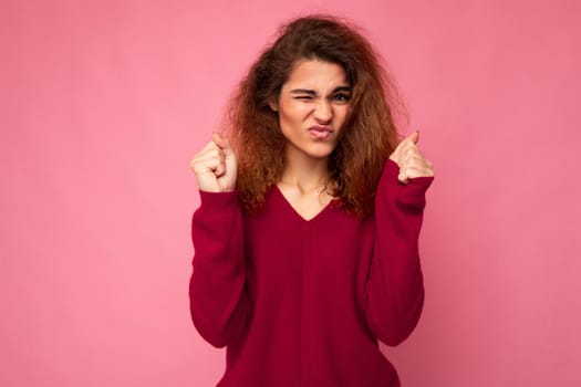 Photo of young emotional attractive brunette curly woman with sincere emotions wearing trendy pink sweater isolated over pink background with empty space and holding fists for good luck. Gesture concept.