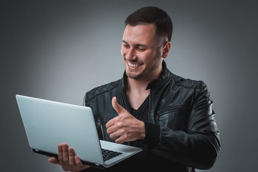 Portrait of young handsome man using laptop, wearing black leather jacket. Studio shot. Emotions