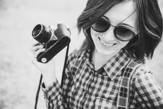 Happy young woman in sunglasses holds retro photo camera. With film grain and low contrast effect. Black-white photo.