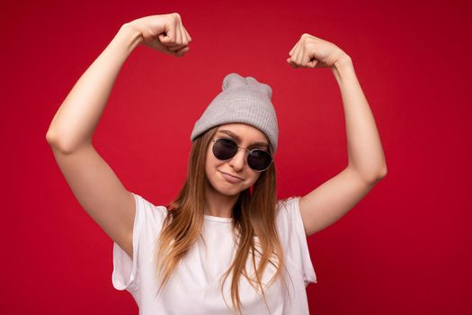 Portrait of young emotional positive funny beautiful dark blond woman wearing casual white t-shirt with empty space for mock up gray hat and sunglasses isolated over red background with free space. Female model showing powerful muscles and biceps. Fitness and workout concept.