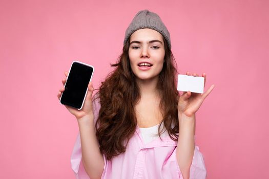 Beautiful happy joyful young curly brunette woman wearing pink shirt and gray hat isolated on pink background with copy space and holding mobile phone with empty screen display and credit card for mockup.