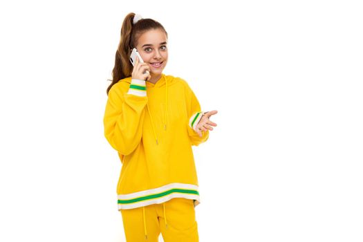 Photo of a beautiful smiling cheerful cute girl with dark hair with a ponytail in a stylish yellow tracksuit with green stripes talking on a mobile phone isolated on white background with free space for text.