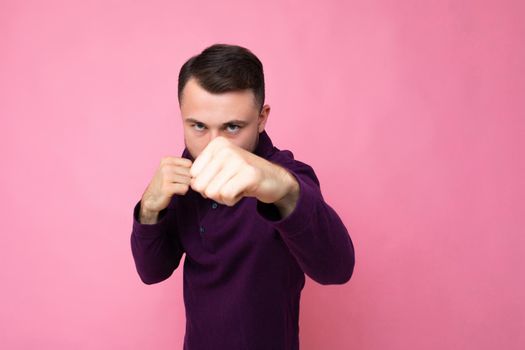 Photo shot of handsome self-confident and good looking young brunet bearded man wearing casual and stylish purple longsleeve poising isolated on pink background with empty space for text. Ready to fight with fist.