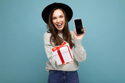 Photo of beautiful positive smiling happy young brunette woman isolated over blue background wall with copy space for text wearing black hat and grey sweater holding white gift box with red ribbon and mobile phone with blank black display screen.
