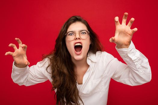 Portrait of young emotional attractive brunette woman with sincere emotions wearing casual white shirt and optical glasses isolated on red background with free space and doing claw gesture as cat with agressive and sexy expression.