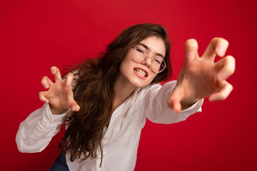 Portrait of young emotional pretty brunette woman wearing casual white shirt and optical glasses isolated on red background with free space and roars doing claw gesture as cat with positive and sexy expression.