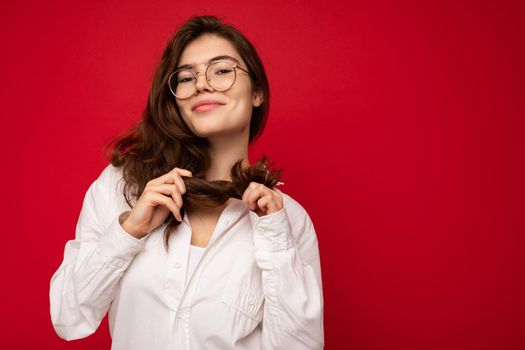 Photo of young beautiful happy smiling brunette woman wearing white shirt and optical glasses. Sexy carefree female person posing isolated near red wall in studio with free space. Positive model with natural makeup.