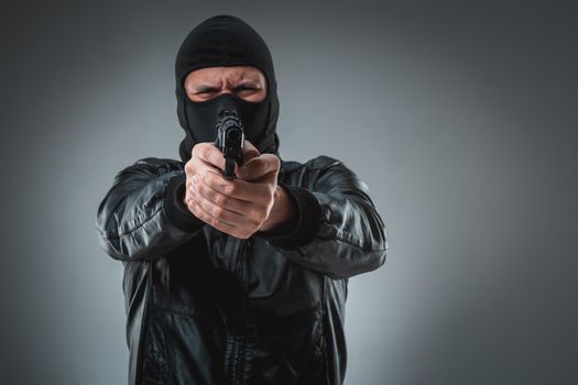 Burglar or terrorist in black mask shooting with gun. A man in the studio on a black-and-gray background