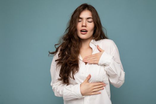 Portrait of young emotional amazed beautiful wave-haired brunette woman with sincere emotions wearing casual white shirt isolated on blue background with empty space and touches chest with hands.