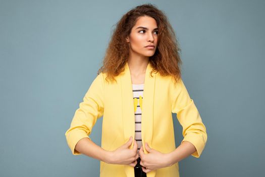 Young self-confident serious beautiful curly brunette woman with sincere emotions poising isolated over background wall with empty space wearing casual yellow jacket. Feelings concept.