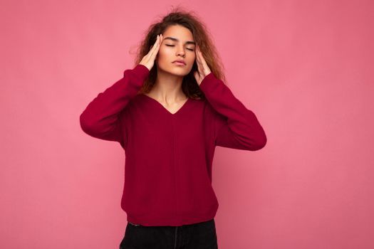 Portrait of young emotional beautiful brunette curly woman with sincere emotions wearing trendy pink pullover isolated on pink background with copy space and having headache.