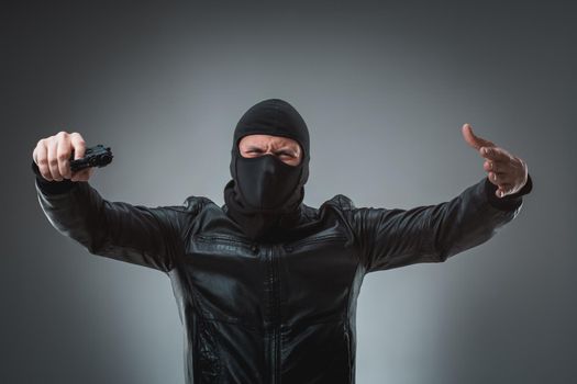 Masked robber with gun, looking into the camera. Studio shot on gray background. Emotions