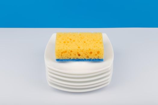 Dishwashing concept, high angle view of cleaning sponge on pile of clean saucers on white table with blue background with copy space. Stack of clean plates and dishwashing sponge close up