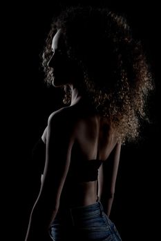 Side lit brunette girl with long curly hair, silhouette studio portrait on dark background.