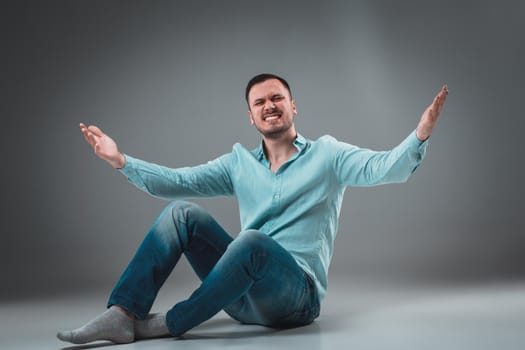 Handsome young man sitting on a floor with raised hands, isolated on gray background. Emotion concept