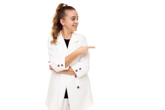 Photo of a beautiful cute adorable smiling brunette teenage girl with a ponytail in a stylish white jacket and white pants isolated on a white background shows a finger on a free space for text.