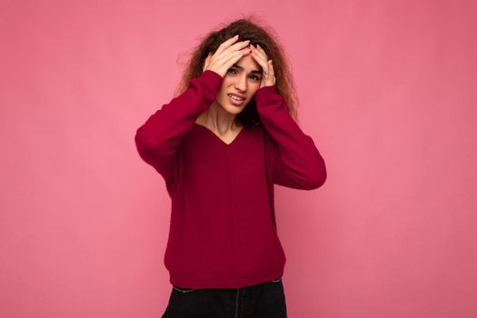 Portrait of young emotional beautiful brunette curly woman with sincere emotions wearing trendy pink pullover isolated on pink background with copy space and having headache.