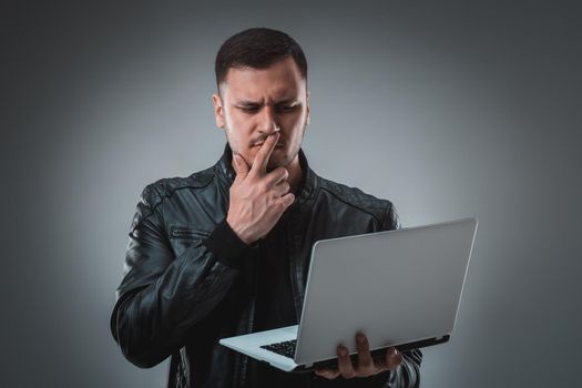 Man in black jacket looking at laptop, half turn. Holding opened laptop and working. Emotion. Indoors, studio, waist up, profile