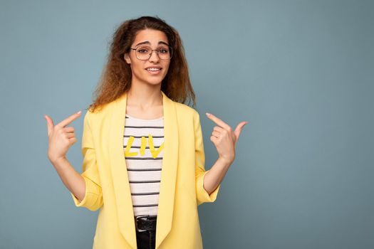 Photo of young positive beautiful brunette curly woman with sincere emotions wearing casual yellow jacket and optical glasses isolated on blue background with copy space and asking pointing at yourself.