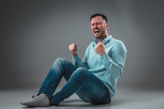 The man is sitting on the floor, isolated on gray background. Man showing different emotions. A man is dressed in blue jeans and a shirt