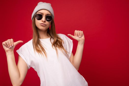 Photo of young positive cool attractive dark blonde woman with sincere emotions wearing casual white t-shirt with empty space for mockup gray hat and sunglasses isolated on red background with copy space and pointing at herself.