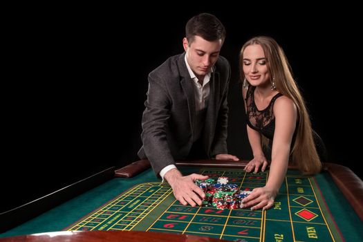 Young beautiful couple takes their winnings at the roulette table at the casino, on a black background. A man in a suit with a woman in a black dress