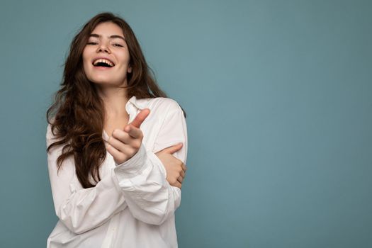 Photo of young positive happy smiling beautiful woman with sincere emotions wearing stylish clothes isolated over background with copy space and pointing at camera.