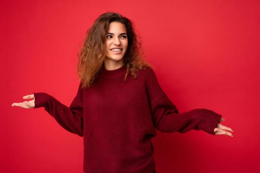 Adult stylish pretty curly brunette female person with sincere emotions poising isolated on background wall with copy space wearing casual dark red sweater. Positive concept.