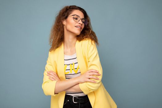 Photo of young positive arrogant beautiful brunette curly woman with sincere emotions wearing casual yellow jacket and optical glasses isolated on blue background with copy space.