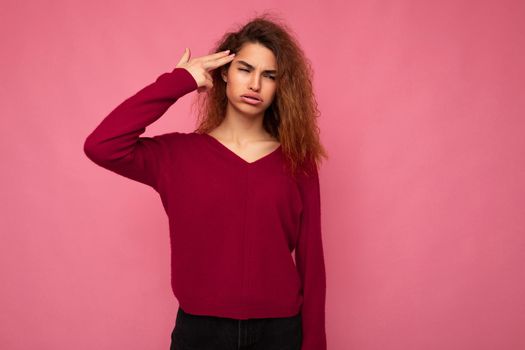 Portrait of young emotional beautiful brunette curly woman with sincere emotions and grimace wearing trendy pink pullover isolated on pink background with copy space and showing gun gesture with hand.