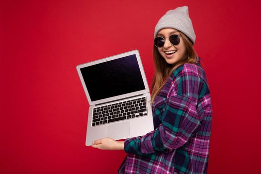 Photo of beautiful pretty attractive happy positive smiling young dark blonde woman isolated over red background with copy space wearing purple stylish shirt, gray hat and casual white t-shirt and sunglasses. Computer laptop with blank black display monitor for mockup.