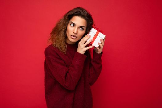 Photo shot of attractive serious thoughtful concentrated young brunette curly woman isolated over red background wall wearing red sweater holding gift box looking to the side and thinking.