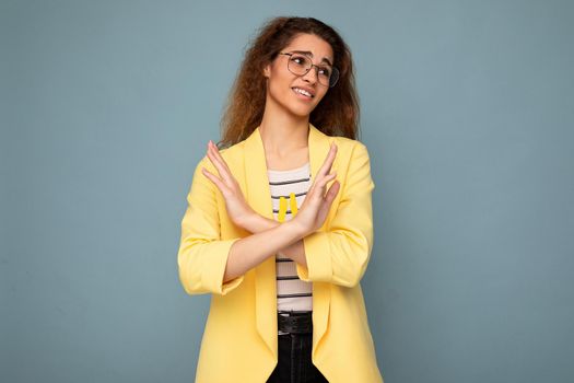 Photo of young emotional attractive brunette curly woman with sincere emotions wearing stylish yellow jacket and optical glasses isolated on blue background with empty space and saying I don't want it.