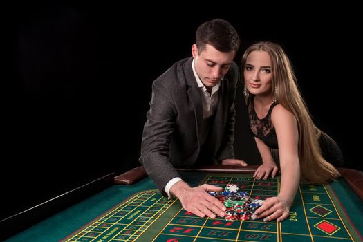 Young beautiful couple takes their winnings at the roulette table at the casino, on a black background. A man in a suit with a woman in a black dress