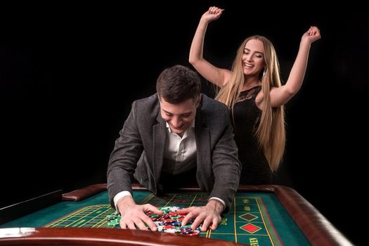 Young beautiful couple takes their winnings at the roulette table at the casino, on a black background. A man in a suit with a woman in a black dress