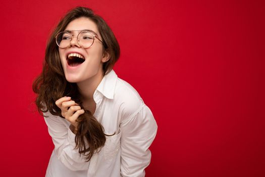 Beautiful young happy joyful funny curly brunette woman wearing white shirt and optical glasses isolated on red background with copy space. Emotional concept
