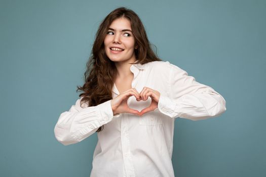 Portrait of young positive happy attractive curly brunette woman with sincere emotions wearing casual white shirt isolated on blue background with free space and making heart form with hands. Love concept.